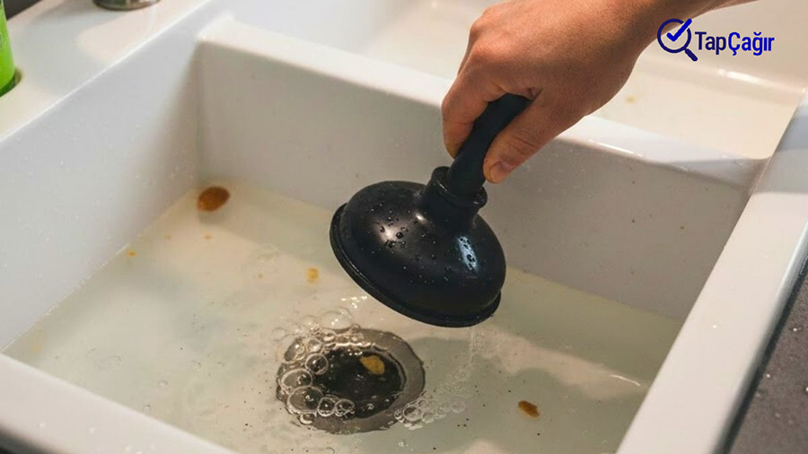 Bir şəxsin əli qara rezin vantuzu tutaraq, içi köpüklü çirkli su və qida qalıqları ilə dolu ikili ağ mətbəx çanağının su axarı üzərinə doğru aparır. Vantuzdan su damcılayır. Şəklin sağ üst küncündə göy rəngli TapÇağır loqosu yer alır